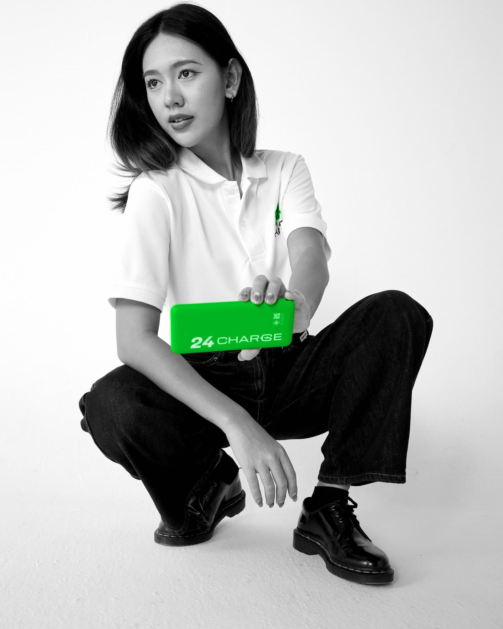 Black-and-white photo with only the product in colour: a woman crouching in a white polo shirt, holding a green 24CHARGE power bank and looking toward the camera