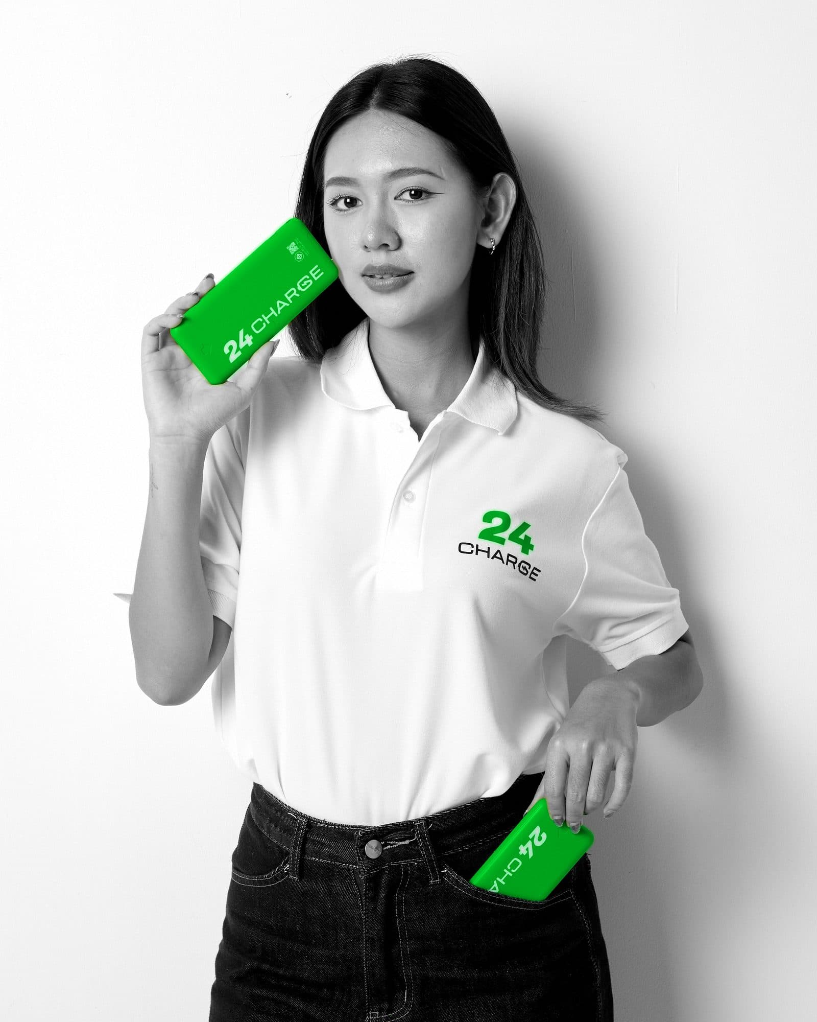 Black-and-white photo with only the product in colour: a woman holding two green 24CHARGE power banks, one by her face and one at her pocket